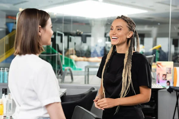 Happy beauty worker welcoming female client, cheerful hair stylist with braids looking at woman in salon, customer satisfaction, beauty industry, hair extension, hair colorist, salon job — Stock Photo