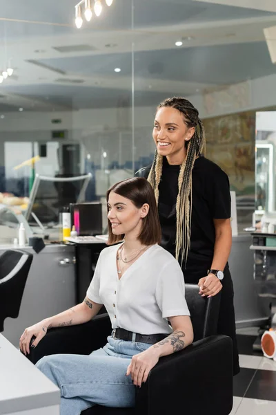 Счастливый парикмахер и клиентка в салоне красоты, веселый работник красоты с косичками стоя рядом с татуированной женщиной, обсуждая лечение волос, наращивание волос, удовлетворенность клиентов — стоковое фото