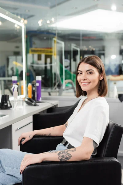 Счастливая клиентка, татуированная молодая женщина сидит в парикмахерском кресле и смотрит на камеру в парикмахерской, услуги красоты, наращивание волос, окрашивание волос, размытый фон, поколение z — стоковое фото