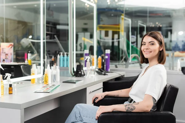 Счастливый клиент салон, татуированная молодая женщина сидит в кресле и смотрит на камеру рядом с инструментами для волос, косметика, наращивание волос, окраска волос, лечение волос, размытый фон, поколение z — стоковое фото