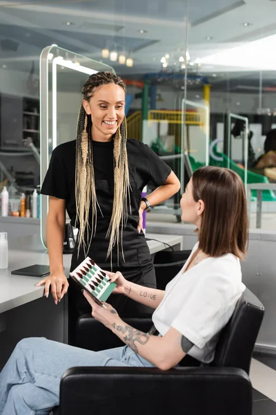 Happy beauty worker standing near female client, holding hair color palette, tattooed woman choosing hair color, customer in salon, hair extension, hair trends, tattoo translation: love — Stock Photo