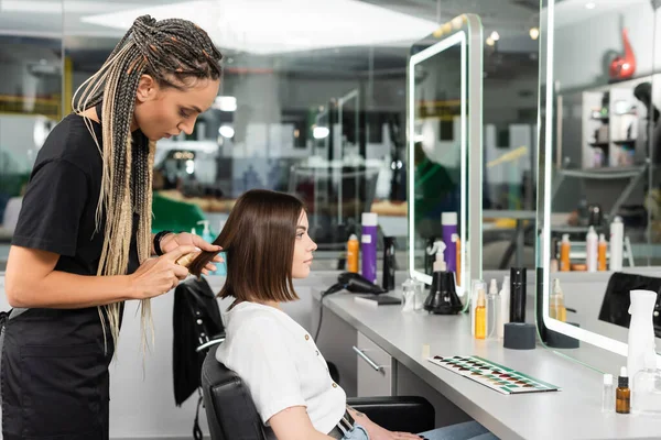 Beauty salon work, professional salon worker with braids applying oil on hair of female client, hairstyling, hair treatment, hairdo, extension, tattooed salon customer, beauty profession, interior — Stock Photo