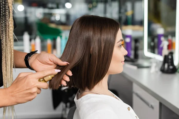 Beauty salon work, professional hair stylist with braids applying oil on hair of woman, hairstyling, hair treatment, hairdo, extension, salon customer, beauty profession, client satisfaction — Stock Photo