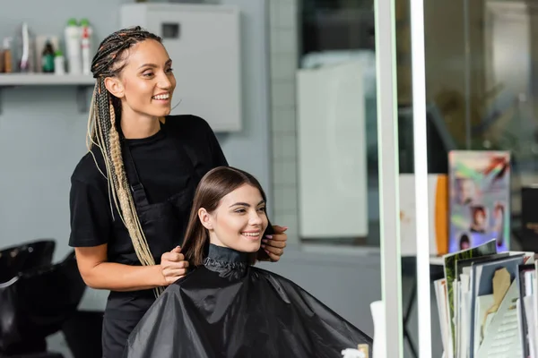 Beauty industry, positivity, happy hair stylist with braids wearing hairdressing cape on female client, hair extension, hair treatment, salon customer, beauty profession — Stock Photo