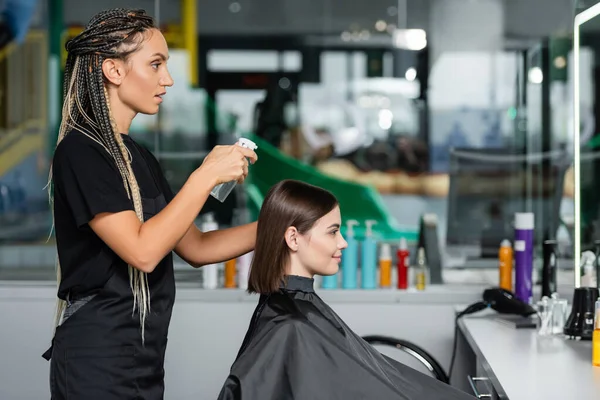 Парикмахер распыляющий волосы клиентки, парикмахер с косичками, держащий аэрозоль рядом с женщиной с короткими волосами брюнетки в салоне, стрижка, лечение волос, парикмахерская, вид сбоку — стоковое фото
