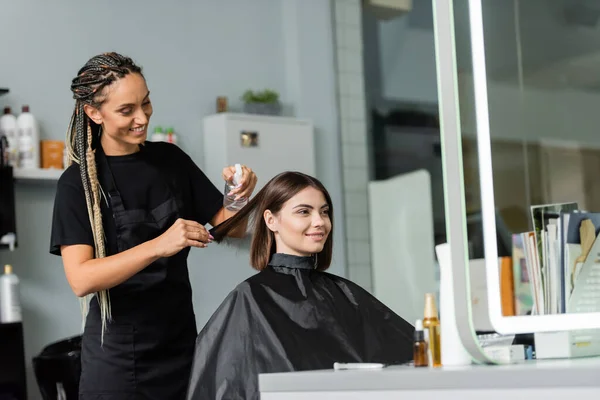 Happy hairstylist spraying hair of female client, hairdresser with braids holding spray bottle near woman with short brunette hair in salon, haircut, hair treatment, hair make over, hairdo — Stock Photo