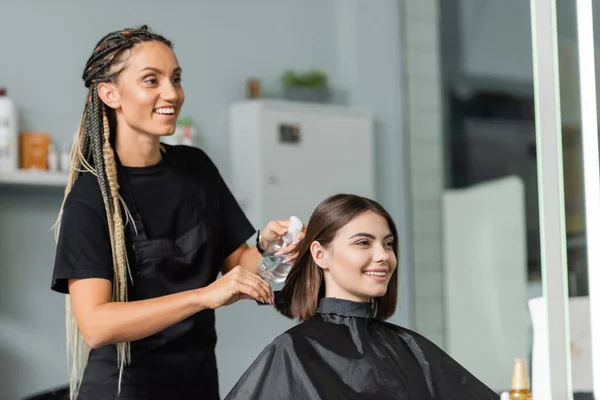 Hairstylist spraying hair of happy woman, hairdresser with braids holding spray bottle near female client with short brunette hair in salon, hair treatment, hair make over, hairdo — Stock Photo