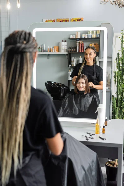 Hairstylist spraying hair of happy woman, hairdresser with braids holding spray bottle near female client with short brunette hair in salon, hair extension, hair treatment, hairdo, blurred foreground — Stock Photo