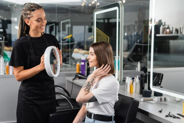 Послуги салону, перукарня з косами тримає дзеркало біля татуйованого жіночого клієнта, щаслива брюнетка жінка з коротким волоссям, салон краси, зачіска, професіонал з волосся, салон краси, інтер'єр — стокове фото
