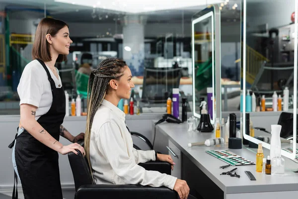 Hair professional, happy and tattooed beauty worker in apron and female client with braids looking at mirror in salon, beauty industry, salon job, customer in salon, hairdresser, side view — Stock Photo