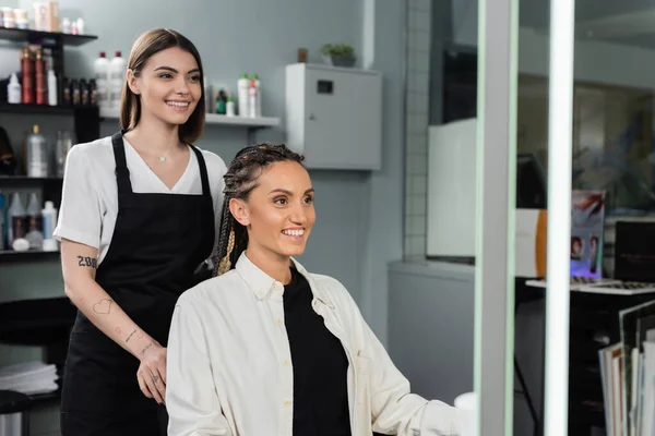 Hair professional, happy and tattooed beauty worker in apron and female client with braids smiling in salon, beauty industry, salon job, customer in salon, hairdresser, salon services — Stock Photo