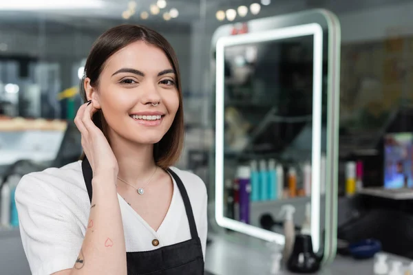 Professional headshots, tattooed beauty worker in apron looking at camera in beauty salon, hair extension, hair stylist, hair coloring, salon job, beauty salon work, hair trends, nose piercing — Stock Photo