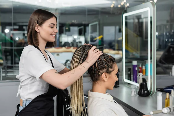 Hair make over, cheerful hair stylist lifting hair of female client with braids, salon customer, beauty profession, client satisfaction, hair fashion, hairdo, tattooed, beauty profession — Stock Photo