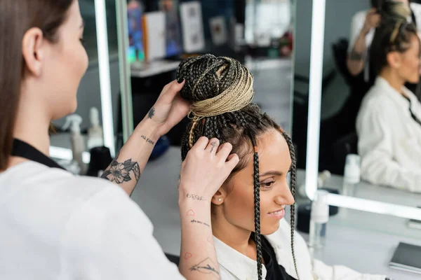 Salon experience, tattooed hairdresser doing hair bun to female client with braids, cheerful women, client satisfaction, customer in salon, beauty service, feminine, hair make over, blurred — Stock Photo