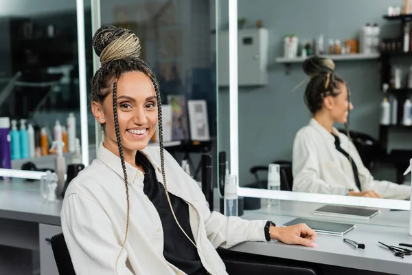 Happy client in beauty salon, cheerful woman with hair bun looking at camera, customer satisfaction, hair salon, hairstyle, female client with braids, looking at camera, mirror refection — Stock Photo