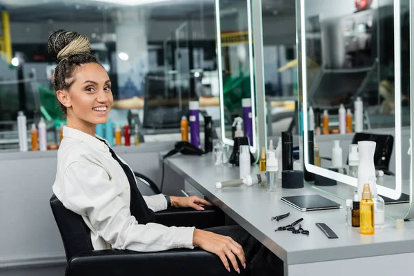 Happy client in beauty salon, cheerful woman with hair bun looking at camera, customer satisfaction, beauty tools, hairstyle, female client with braids, mirror refection — Stock Photo
