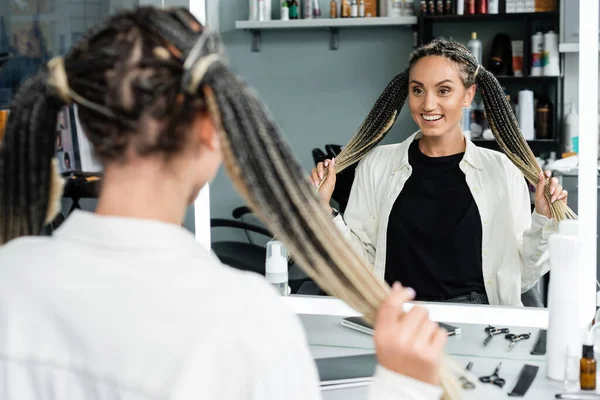 Щасливий клієнт в салоні краси, весела жінка з косами дивиться на дзеркало, задоволення клієнтів, салон краси, зачіска, жіночий клієнт з косами, дзеркальна рефлексія, два хвости, розширення — стокове фото