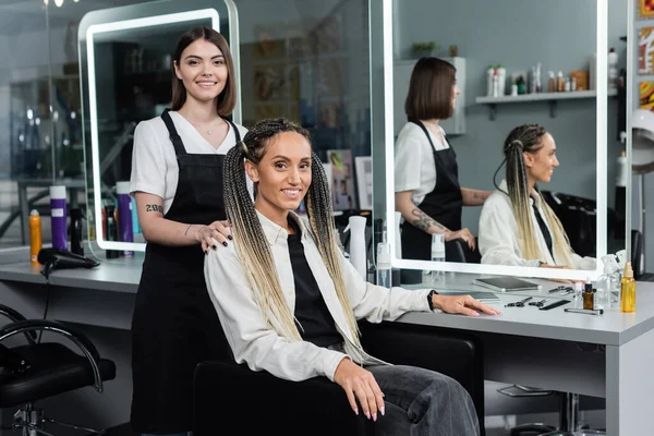 Hairdresser and female client, beauty salon, cheerful woman with braids and two ponytails smiling near hair stylist in salon, customer satisfaction, beauty worker, professional, hair fashion — Stock Photo