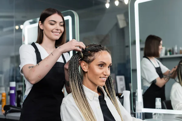 Hairdresser and female client, beauty salon, tattooed hair stylist doing hair of cheerful woman with braids, two ponytails, customer satisfaction, beauty worker, professional, hair fashion — Stock Photo