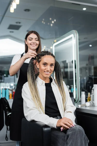 Hairdresser and client, beauty salon, cheerful woman with braids and two ponytails smiling near hair stylist in salon, customer satisfaction, beauty worker, professional, hair fashion — Stock Photo