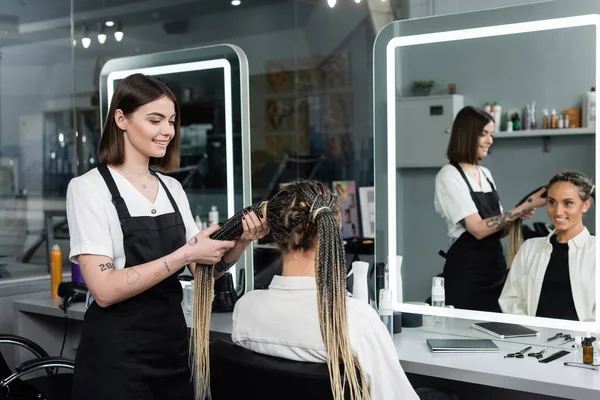 Перукар і клієнт, салон краси, татуйований стиліст робить волосся веселою жінкою з косами, два поні, задоволення клієнтів, працівник краси, мода волосся, дзеркальне відображення — стокове фото