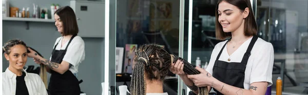 Beauty worker and client, beauty salon, tattooed hair stylist doing hair of cheerful woman with braids, two ponytails, customer satisfaction, beauty worker, hair fashion, mirror reflection, banner — Stock Photo