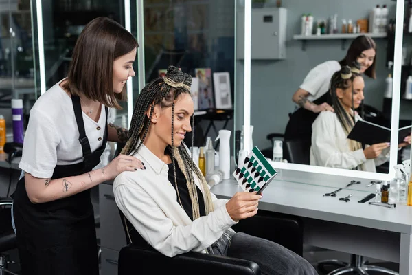 Happy woman in beauty salon, joyful client with braids looking at hair palette near tattooed hairdresser, customer satisfaction, hairstyle, mirror refection, hair buns, braided hair — Stock Photo