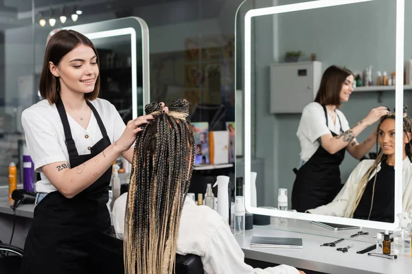 Beauty, hair industry, tattooed hairdresser styling hair of woman with braids, customer satisfaction, hairstyle, mirror reflection, hair buns, braided hair, beauty salon, hair fashion — Stock Photo
