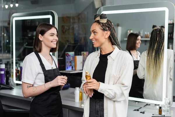 Client satisfaction, hairdresser smiling with female customer, happy woman with braids holding hair oil, hairstyle, hair buns, braided hair, beauty salon, hairstyling product — Stock Photo