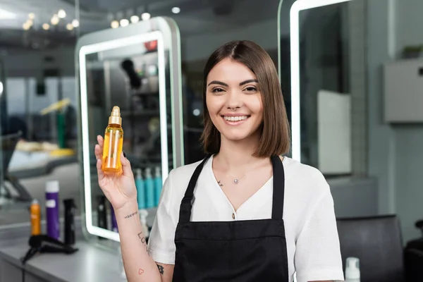 Positivity, tattooed beauty worker in apron looking at camera in beauty salon, suggesting hair oil, advertisement, hair stylist, hair coloring, salon job, beauty salon work, hair product — Stock Photo