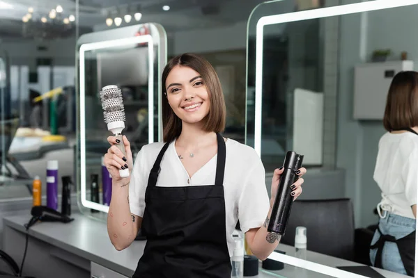 Professional headshots, positivity, tattooed hairdresser in apron holding round brush and bottle with hair spray, beauty worker, looking at camera, smiling, beauty salon, salon worker — Stock Photo