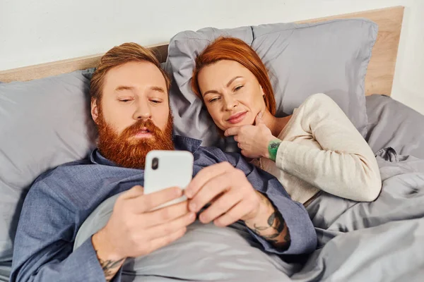 Рыжая пара проводит время без детей, выходной, муж и жена, бородатый мужчина с помощью смартфона рядом с женщиной, удобная гостиная, уютная спальня, беззаботный, татуировка — стоковое фото