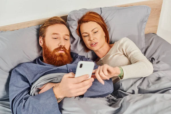 Tattooed couple spending time without kids, day off, husband and wife, bearded man using smartphone near redhead woman, comfortable living, cozy bedroom, carefree, screen time — Stock Photo