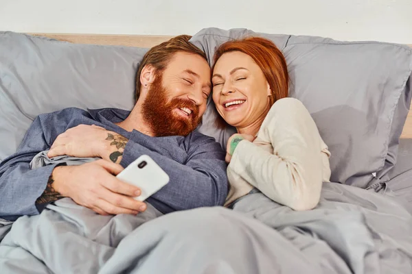 Carefree, laughter, screen time, tattooed couple spending time without kids, day off, husband and wife, bearded man and redhead woman, smartphone users, cozy bedroom, home environment — Stock Photo