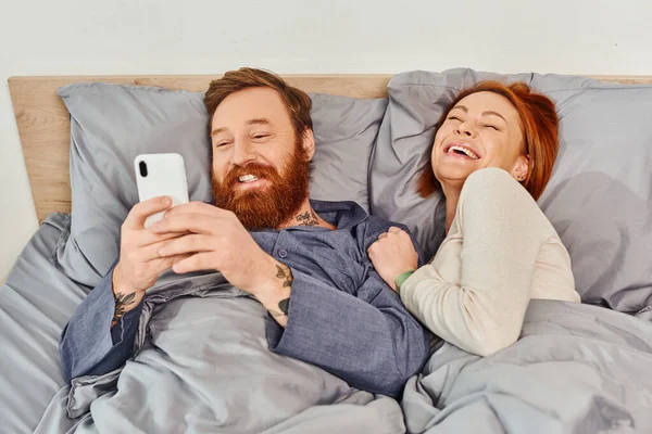 Смех, время релаксации, татуированная пара без детей, муж и жена, бородатый мужчина с помощью смартфона рядом с рыжей женщиной, уютная спальня, беззаботный, экран время, выходной — стоковое фото