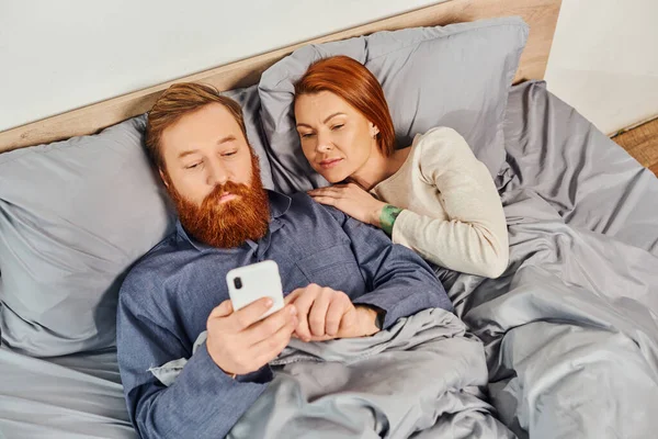 Networking, relaxation time, tattooed couple relaxing, weekends without kids, husband and wife, bearded man using smartphone near redhead woman, cozy bedroom, screen time, day off, top view — Stock Photo