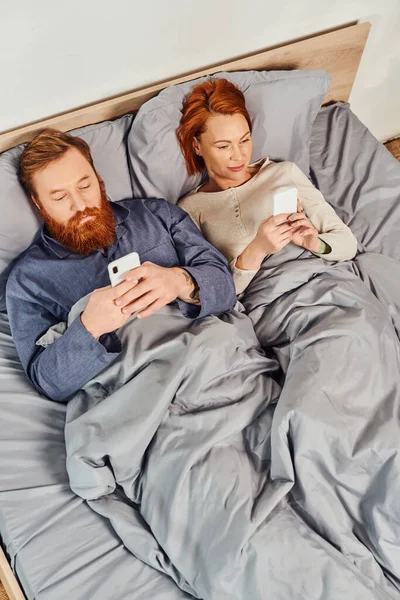 Вид сверху, татуированная пара с помощью смартфонов, общение, отдых по выходным без детей, муж и жена, бородатый мужчина с помощью смартфона рядом с рыжей женщиной, уютная спальня, время на экране — стоковое фото
