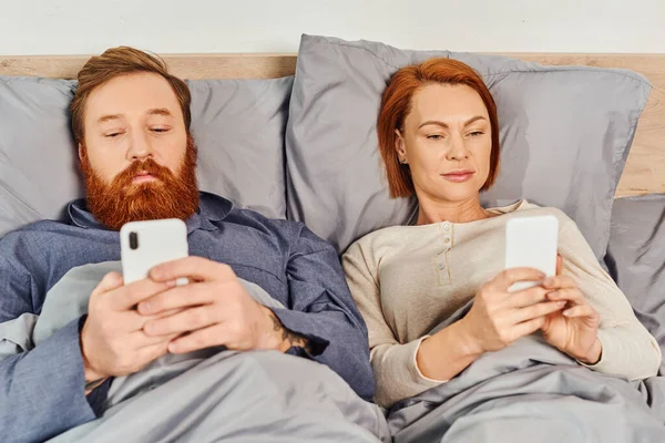 Татуированная пара с использованием смартфонов, сетей, отдыха в выходные дни без детей, муж и жена, бородатый мужчина и рыжая женщина с мобильными телефонами, уютная спальня, экран — стоковое фото