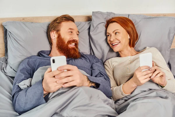 Батьки вдома, татуйована пара, використовуючи смартфони, мережу, розслабляючись на вихідних без дітей, щасливий чоловік і дружина, затишна спальня, час екрану — стокове фото