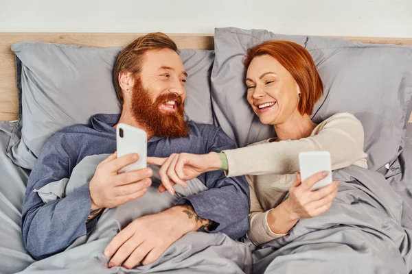 Час екрану, татуйована пара використовує смартфони, мережу, розслабляється на вихідних без дітей, чоловіка та дружини, руда жінка вказує на мобільний телефон бородатого чоловіка, затишну спальню — стокове фото