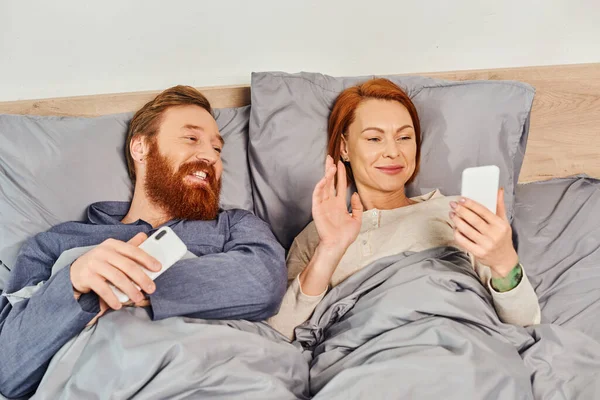 Экранное время, татуированная пара с помощью смартфонов, расслабляющая в выходные без детей, муж и жена, рыжая женщина с видеозвонком рядом с бородатым мужчиной, уютная спальня, машущая рукой — стоковое фото