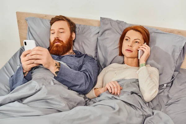 Татуированная пара с помощью смартфонов, бородатый мужчина в сети, расслабляющий в выходные, муж и жена, рыжая женщина разговаривает с детьми на мобильных телефонах, уютная спальня, время на экране — стоковое фото