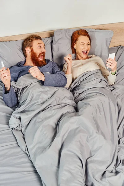 Tattooed couple using smartphones, networking, relaxing on weekends without kids, excited husband and wife, bearded man and redhead woman with mobile phones, cozy bedroom, screen time — Stock Photo
