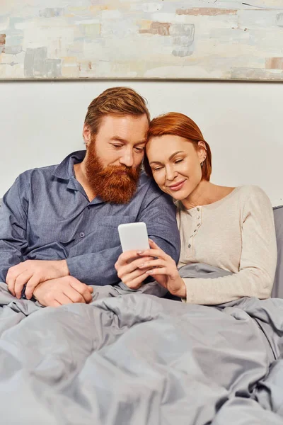 Цифровая пара, экранное время, общение, отдых по выходным без детей, довольные муж и жена, бородатый мужчина и рыжая женщина с помощью смартфона, татуировка, выходной — стоковое фото