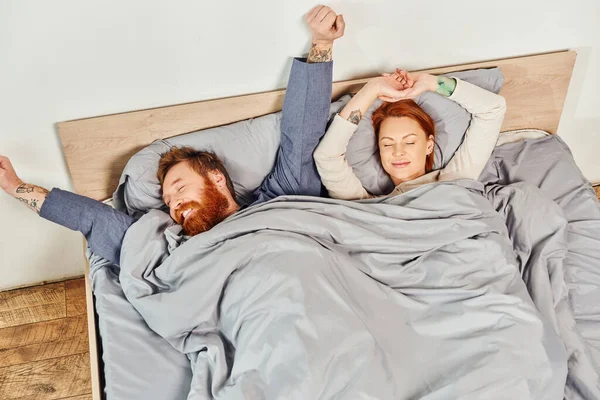 Quiet house, parents alone at home, redhead husband and wife in cozy bedroom, bearded man and carefree woman relaxing on weekends, day off, wake up, tattooed, stretching, closed eyes, top view — Stock Photo