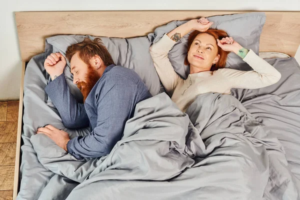Quiet house, parents alone at home, redhead husband and wife in cozy bedroom, bearded man sleeping near carefree woman relaxing on weekends, day off, wake up, tattooed, enjoy, top view — Stock Photo