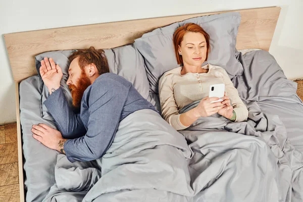 Quiet house, parents alone at home, redhead husband and wife in cozy bedroom, bearded man sleeping near carefree woman using smartphone, networking, screen time, day off, tattooed — Stock Photo