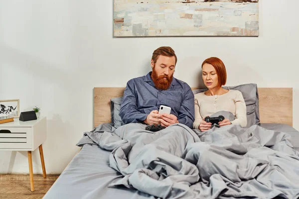 Quiet house, couple without kids, redhead husband and wife, bearded man using smartphone near woman holding joystick, networking, playing video game, day off, tattooed, parents alone at home — Stock Photo