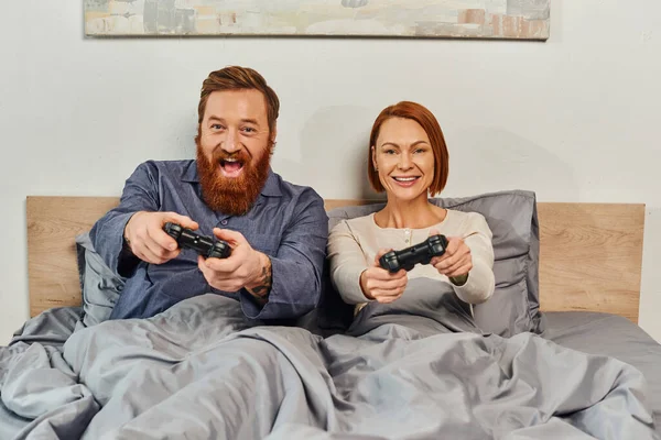 Fins de semana sem filhos, marido e mulher ruiva jogando videogame, homem barbudo e mulher feliz segurando joysticks, animado, diversão de jogos, casal, estilo de vida moderno — Fotografia de Stock