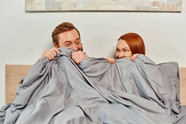 Day off without kids, redhead husband and wife looking at each other, hiding behind blanket, married couple, modern lifestyle, happiness at home, relaxation time, parents alone at home — Stock Photo
